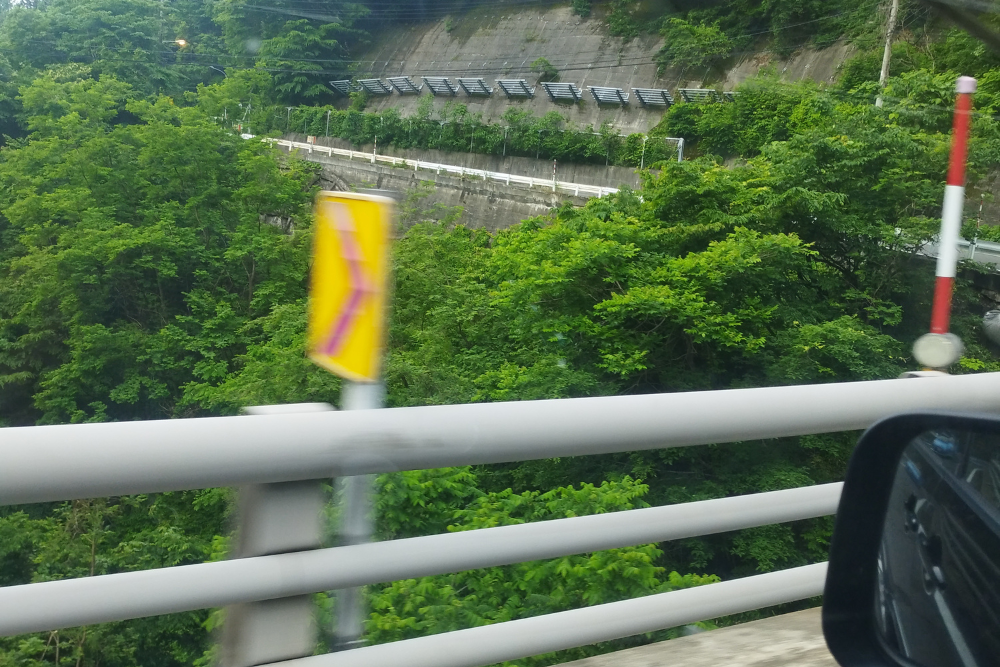 「車で山道を走行中に撮影した、緑に囲まれた山の斜面と道路」