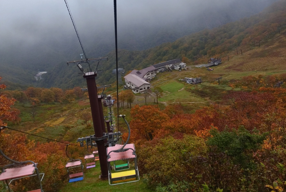 「紅葉した山の斜面を進むリフトから見た山小屋と色づいた木々」